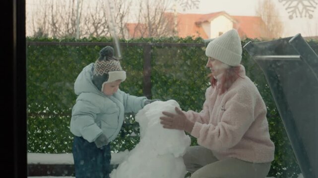 A mother and her young son work together to create a snowman in the yard of their apartment building on a snowy day. They enjoy their time playing in the snow.