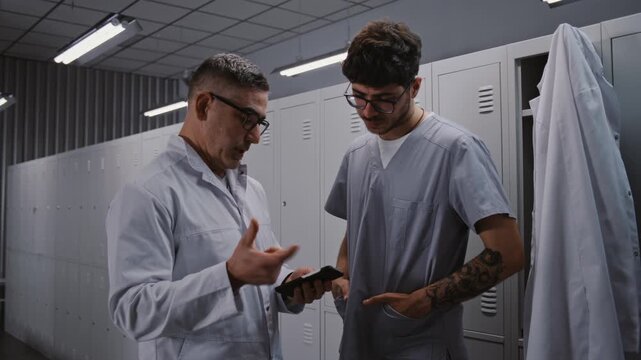 After Busy Workday, Two Male Doctors Relax by Grey Metal Lockers, Viewing Sport News on Smartphone and Chatting About Personal Interests in Locker Room. Concept of Shift End and Employee Wellness.