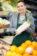 Obraz premium Neat woman selling radishes on a shelf in a supermarket. Farm shop owner offering organic radishes and vegetables to customers