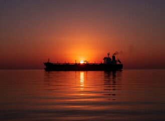 Fototapeta premium Crude oil tanker sailing across calm waters at vibrant orange sunset with reflection