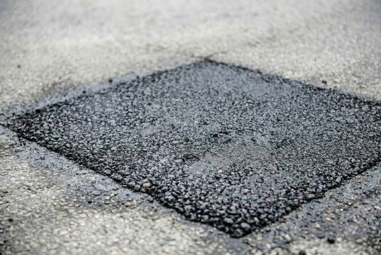Close-up of a freshly patched asphalt road surface with a diamond-shaped repair, showing texture and wetness.