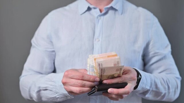 A person is sorting through cash in a wallet while standing against a plain background. Close-up of hands counting Russian rubles