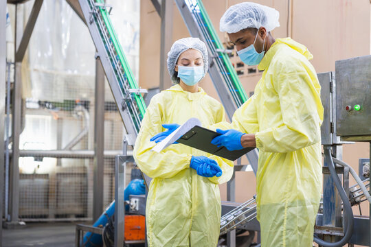 Diverse factory workers in yellow PPE suits checking production data with clipboard  in modern industrial facility, Quality control inspector wearing masks and hairnets working in food or pharma plant