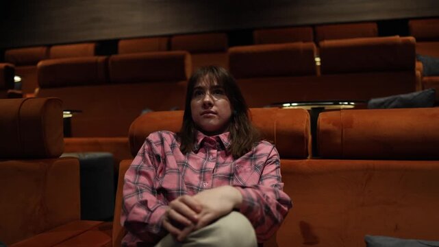 Young woman sits in empty cinema hall watching film with thoughtful expression soft light on face quiet atmosphere and immersive movie experience