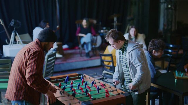 Two young men play foosball together in a casual setting. Other students socialize and enjoy games in the background.