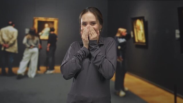Woman covers mouth with hands in an art museum building while viewing framed paintings and nearby visitors; surprise wonder.