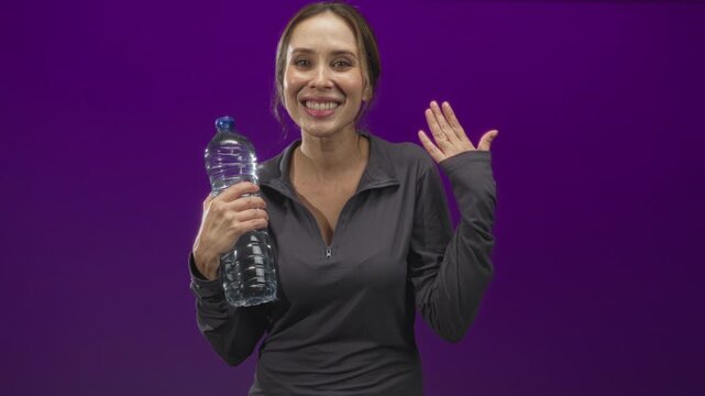 Woman holding a large plastic water bottle and waving right hand in studio with purple backdrop, smiling at camera; hydration joy.