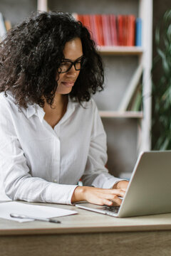 A woman with curly hair and glasses is typing on a laptop while a camera is set up in front of her, suggesting she is creating video content