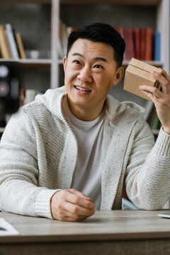An Asian man sits at a desk, holding a small cardboard box while looking into a camera recording him for a video