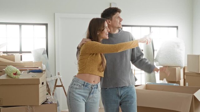 Man and woman embrace with arm around shoulder amid packed boxes and paint roller, smiling and pointing toward window in building; moving day joy.