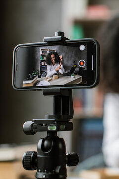 A young woman with curly hair is recording a video blog or tutorial using her smartphone mounted on a tripod, showcasing a product