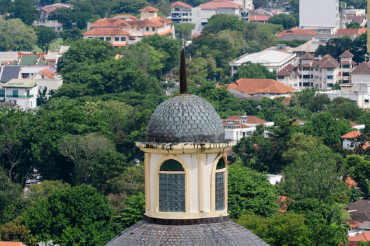 Close up of an old domed tower with textured roof tiles and arched windows, set against a backdrop of lush greenery and urban residential buildings.