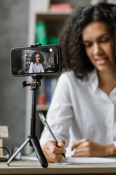 A young woman with curly hair is filming herself with a smartphone on a tripod while taking notes for an online lesson