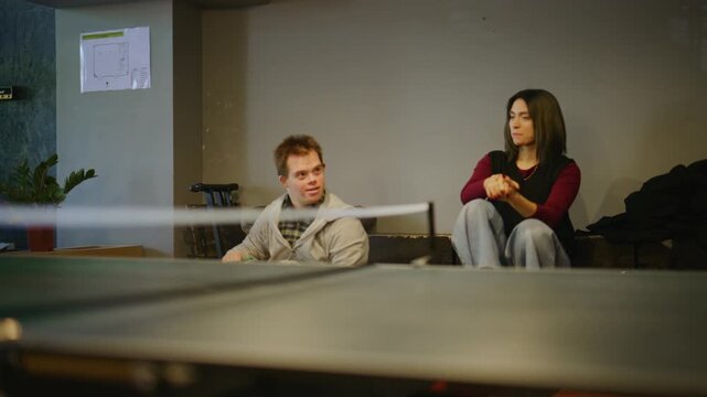 A young man with Down syndrome and a woman watch a ping-pong game. They react with smiles and supportive gestures.
