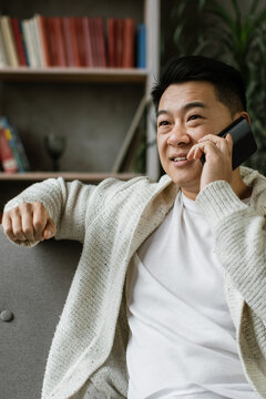 A middle-aged Asian man in a cozy cardigan is casually talking on his smartphone while relaxing on a sofa in his living room