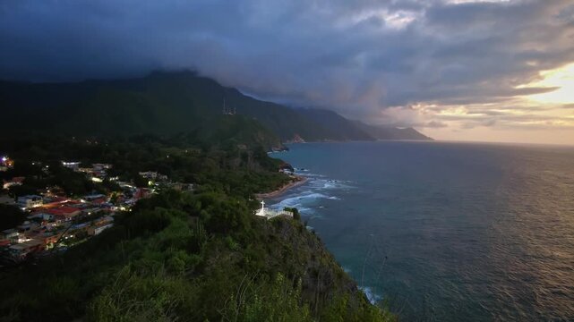 Handheld aerial view of the bay in the beach town of Choroni, with dense mountains and a cloudy horizon at sunset, Aragua, Venezuela