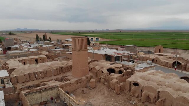  Aerial view of ancient Persian city ruins with historic mud brick structures, windcatchers, and old alleys, surrounded by green fields under cloudy sky in Yazd, Iran. 