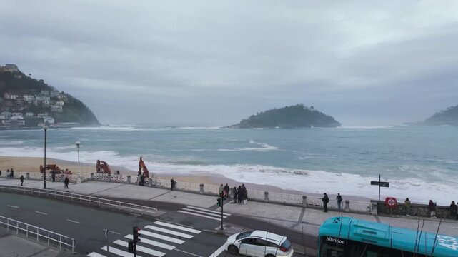 Scenic view of Ondarreta beach in San Sebastian on a cloudy day, with people on the promenade watching rough sea waves crashing on the sand and cars driving on the waterfront street