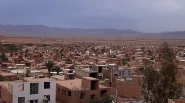  Expansive panoramic view of Yazd city, Iran, with old neighborhoods, mudbrick structures, brown rooftops, and distant arid mountains under a cloudy sky. Persian architectural style. 