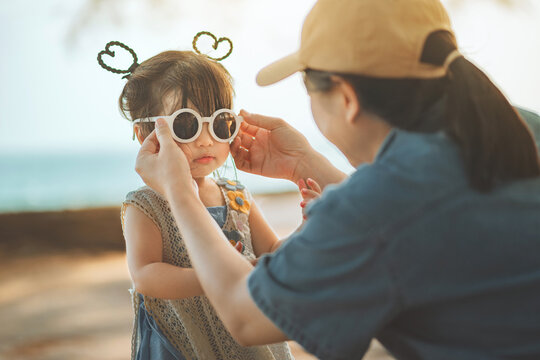 Caring Asian mother putting sunglasses on her toddler daughter at an outdoor beach park. Lifestyle scene captures family bonding and sun protection during a summer travel holiday.