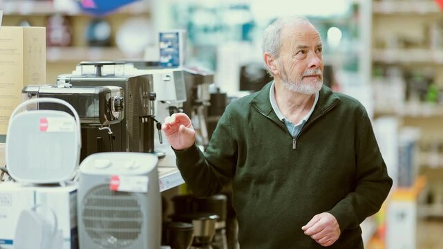 Senior man buyer examines coffee machine and decides on purchase. Client in hardware store picks up household appliances. Store of household appliances and household goods, showroom