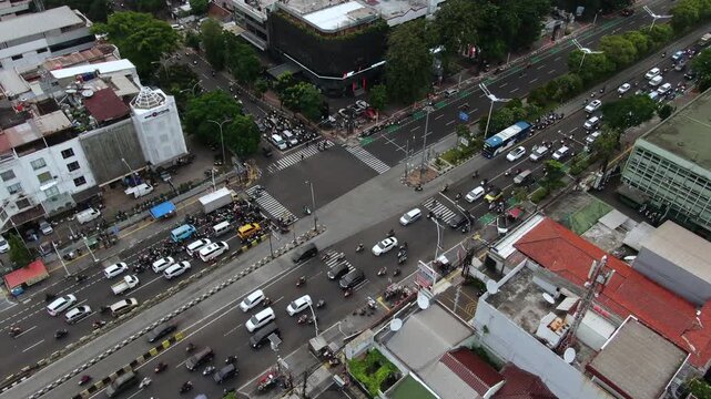 Aerial High Angle View of Busy City Intersection with Heavy Traffic. Featuring TransJakarta Buses, Cars, and Motorcycles Navigating Urban Roads Surrounded by Commercial Buildings and Residential Areas