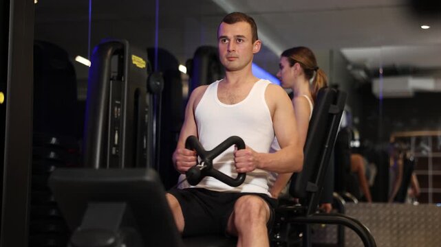 Determined young guy training his arms pulling handle of cable exercise machine in fitness hall. High quality 4k footage