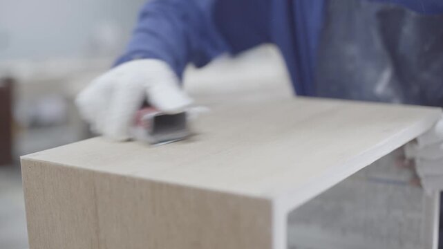 Craftsman wearing white protective gloves while meticulously sanding the edge of a light-colored wooden furniture piece. 