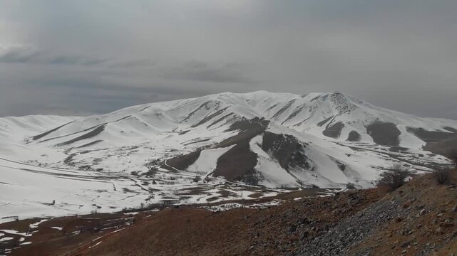  Snow capped mountains dominate a vast winter landscape with rolling hills and a winding road, showcasing the serene beauty of a cold, natural environment under a cloudy sky. 