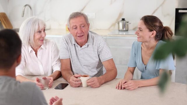 Smiling elderly parents and adult children sit at the table with tea and talk about home life