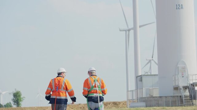 Back of two Caucasian engineers or technicians work in front of cluster of windmill or wind turbine and walk to it for maintenance or manage system in workplace.