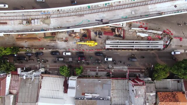 Aerial top down view of elevated railway construction project. Top angle shot of infrastructure development featuring railway tracks, heavy machinery, and busy city traffic flow on the urban road