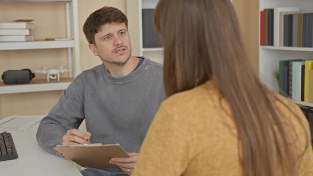 Man taking notes on a clipboard while listening intently to woman seated across a desk in a building; quiet empathy concern.