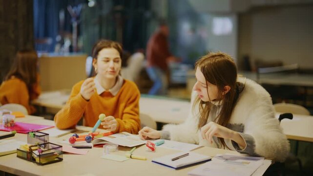 Two female students are working together on a colorful science model at a desk. They are engaged in a collaborative learning activity in a classroom setting.