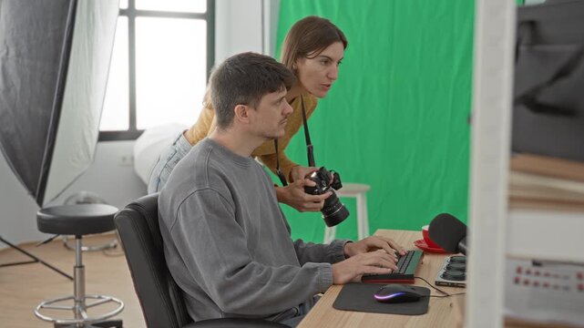 Man typing on keyboard while woman holds camera in studio with green screen; creative teamwork focused.