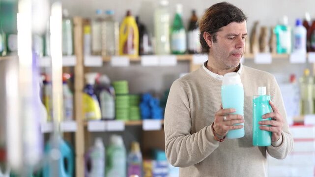 Among shop windows, man buyer leisurely examines goods and chooses shampoo or shower gel. Client reads label, gets acquainted with consumer information