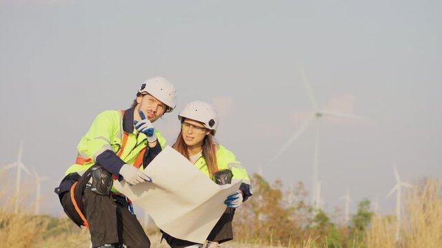 Front view of Caucasian engineer man and woman sit on the ground and discuss together with using drawing paper and stay in front of row of windmill or wind turbine.