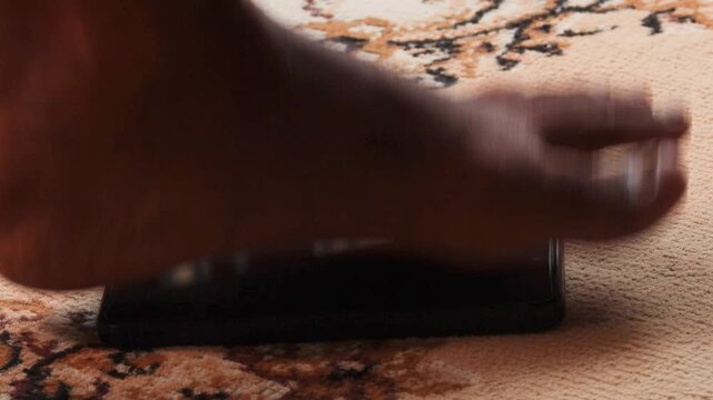 Close-up of anger person bare foot stepping onto smartphone lying on carpeted floor, symbolizing digital detox, breaking addiction, or protesting against technology influence. 