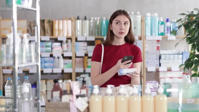 Near display case in pharmacy, young woman scans barcode on ointment for bruises, reads user manual. Client studies product in detail, collects information online.