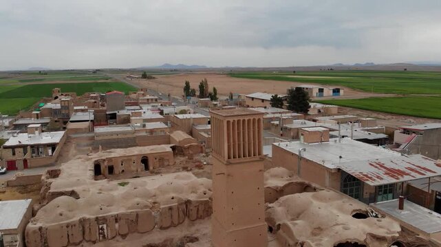  Panoramic rooftop shot of a historical town in Yazd, Iran, with prominent windcatchers and brown adobe homes situated in a desert environment, green fields on the edge, cloudy sky. 