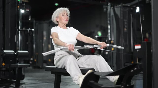 Fit senior woman performing strength training with seated cable row machine in modern fitness facility, focusing on back strength, posture, and staying active for better health