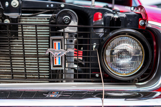 Close-up of a Ford Mustang black radiator grille featuring the iconic running horse emblem with tri-bar, round Sylvania glass headlight, chrome bumper with reflection, visible radiator core and engine