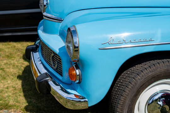 Close-up of the front fender and headlight of a classic light blue FSO Warszawa car, featuring the chrome model name script, metallic bumper, part of the grille, and a tire with a white rim in bright 