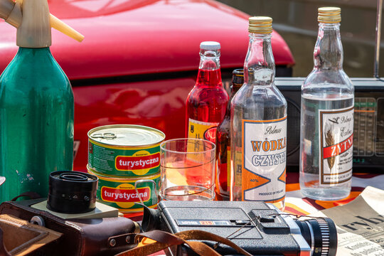 Vintage PRL still life featuring Polish vodka bottles, canned tourist meat, a green soda syphon, retro Lomo video camera, glassware, and old radio set against a red car background.