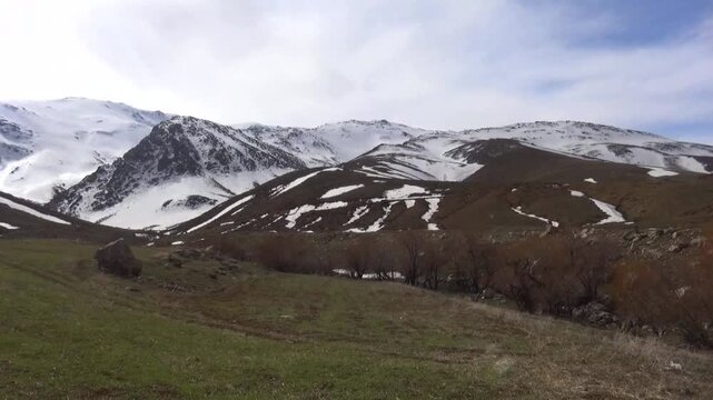  Rolling hills with patches of snow and brown vegetation meet distant, majestic mountain ranges under a clear blue sky, depicting a serene, transitional highland landscape. 