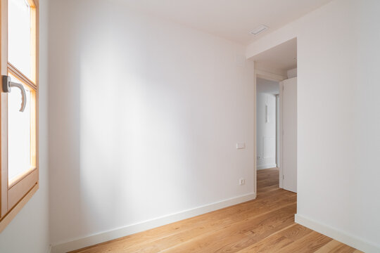 A bright, empty hallway with white walls and wooden flooring. Natural light streams in through a window, creating a spacious and airy atmosphere.