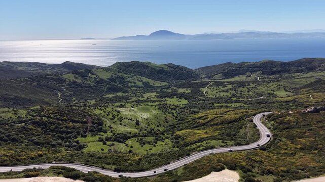 Wide aerial perspective showing a winding coastal road in the foreground with the African coastline visible across the shimmering Strait of Gibraltar.