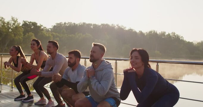 Young happy smiling people doing sport jumping and sits up exercises in nature. Sporty men and women wearing sportswear having workout in park outdoors. Healthy lifestyle. 4k video. Slow motion.