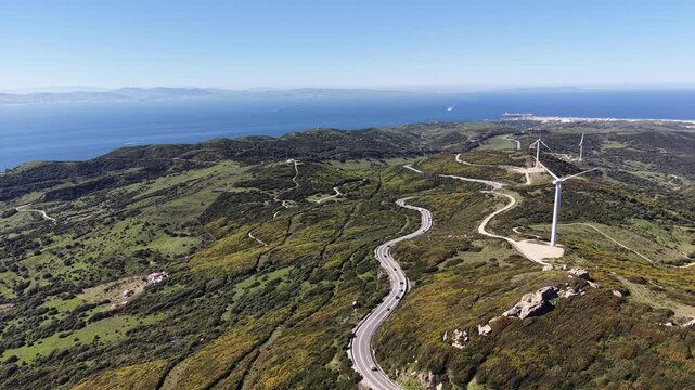 Cinematic aerial shot of massive wind turbines turning on green ridges with a winding road below and the sea stretching toward Africa.