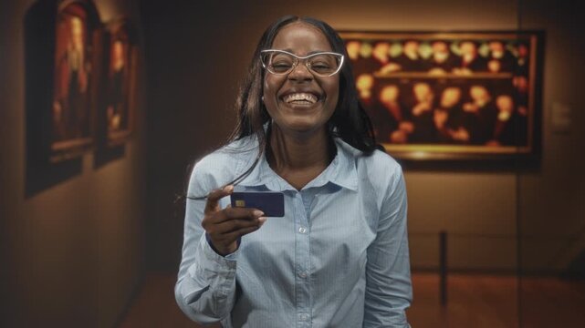 Woman holding creditcard smiling and laughing, wearing glasses and a buttoned shirt in a museum building with framed paintings and stanchion ropes; joy satisfaction.